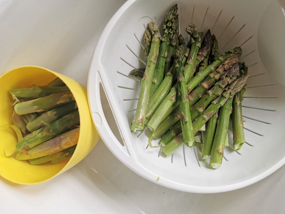 Washed asparagus in white colander and discarded stalks in yellow bowl.