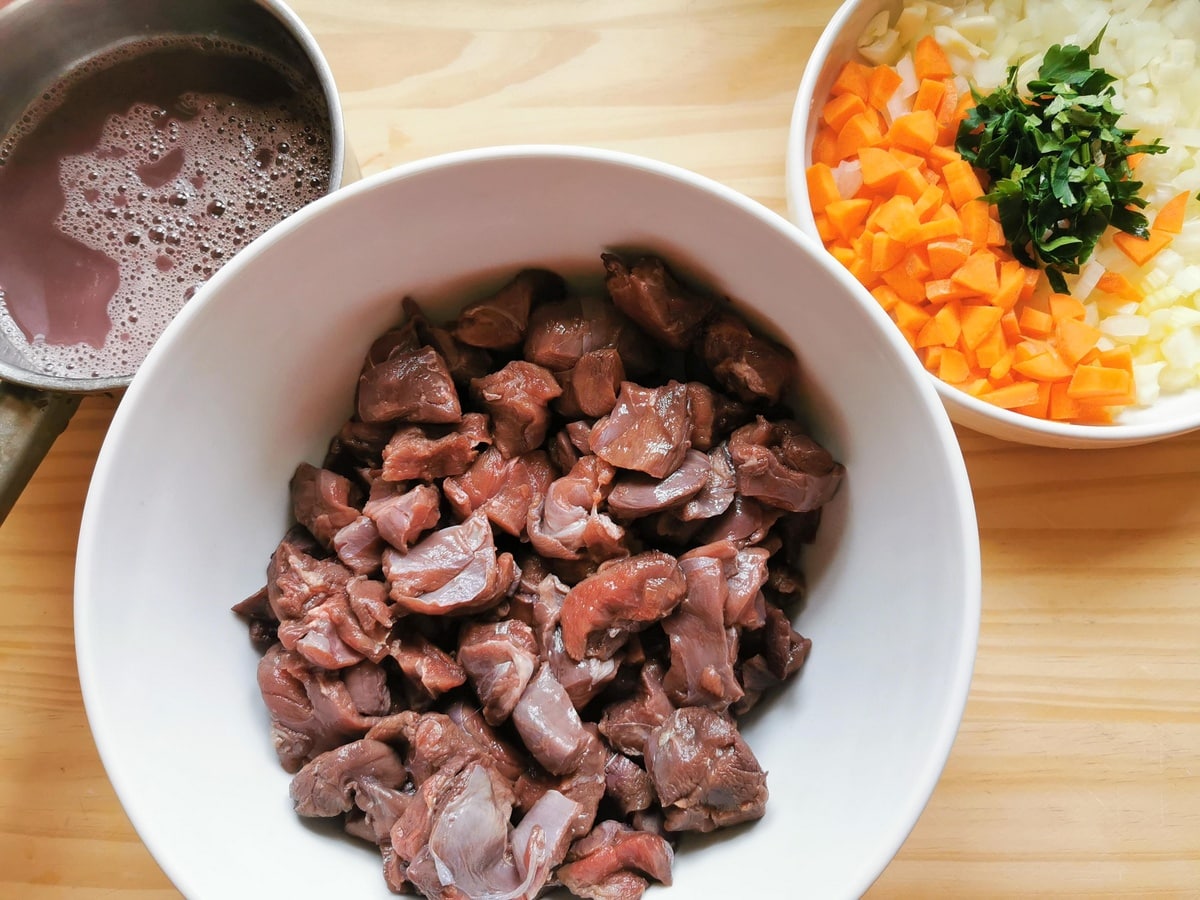 Marinated hare meat pieces in white bowl next to chopped vegetables in white bowl and drain wine marinade in pan.