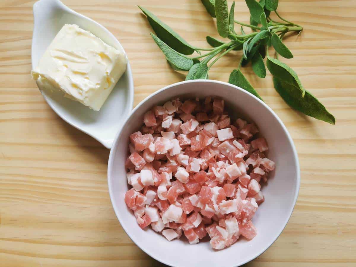 Sage leaves, butter and a bowl of pancetta cubes on wood work surface.