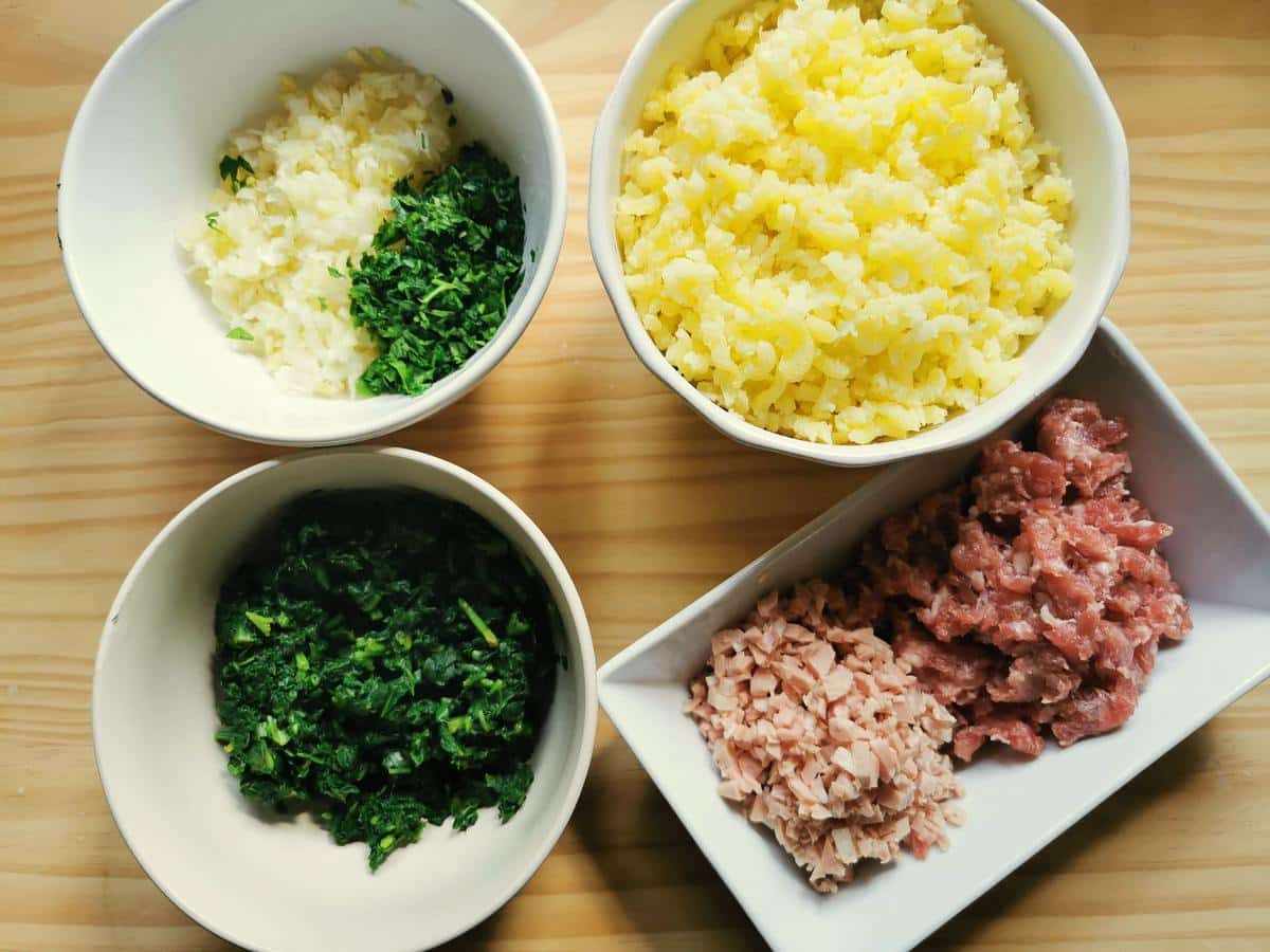 Chopped sausage meat and mortadella in white bowl. boiled and riced potatoes in white bowl. Cooked and chopped spinach in white bowl. Chopped leeks and parsley in white bowl.