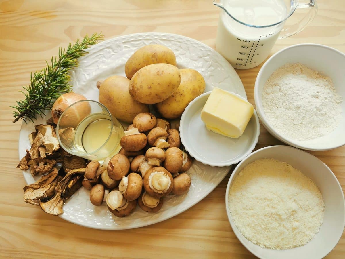 Mushrooms, potatoes, rosemary, butter, flour, milk, shallot and parmesan on a kitchen table.
