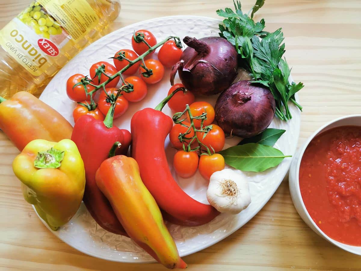 Peppers, onions, garlic, cherry tomatoes, bay leaf, parsley, white wine vinegar and passata on a kitchen table.